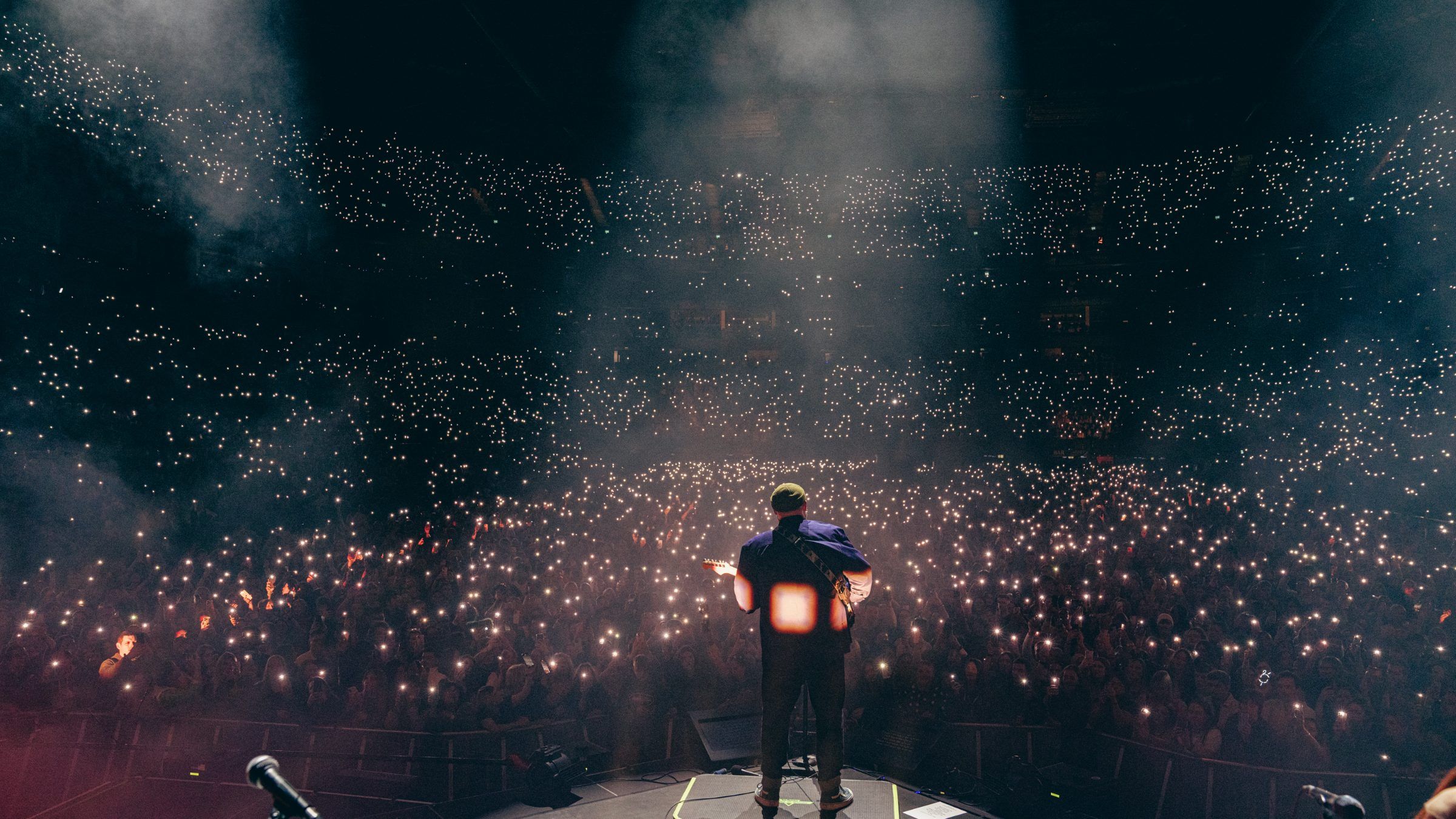 A performer on stage facing an arena crowd lit by thousands of phone lights